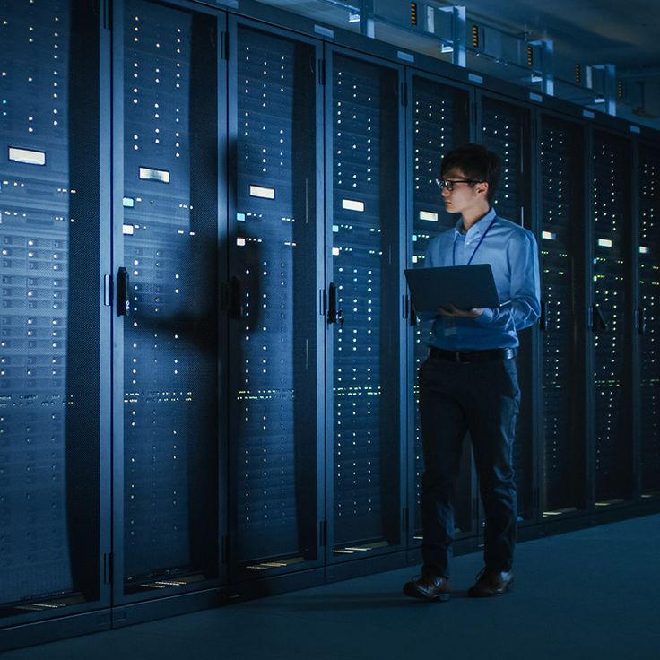 In Dark Data Center: Male IT Specialist Walks along the Row of Operational Server Racks, Uses Laptop for Maintenance. Concept for Cloud Computing, Artificial Intelligence, Supercomputer, Cybersecurity